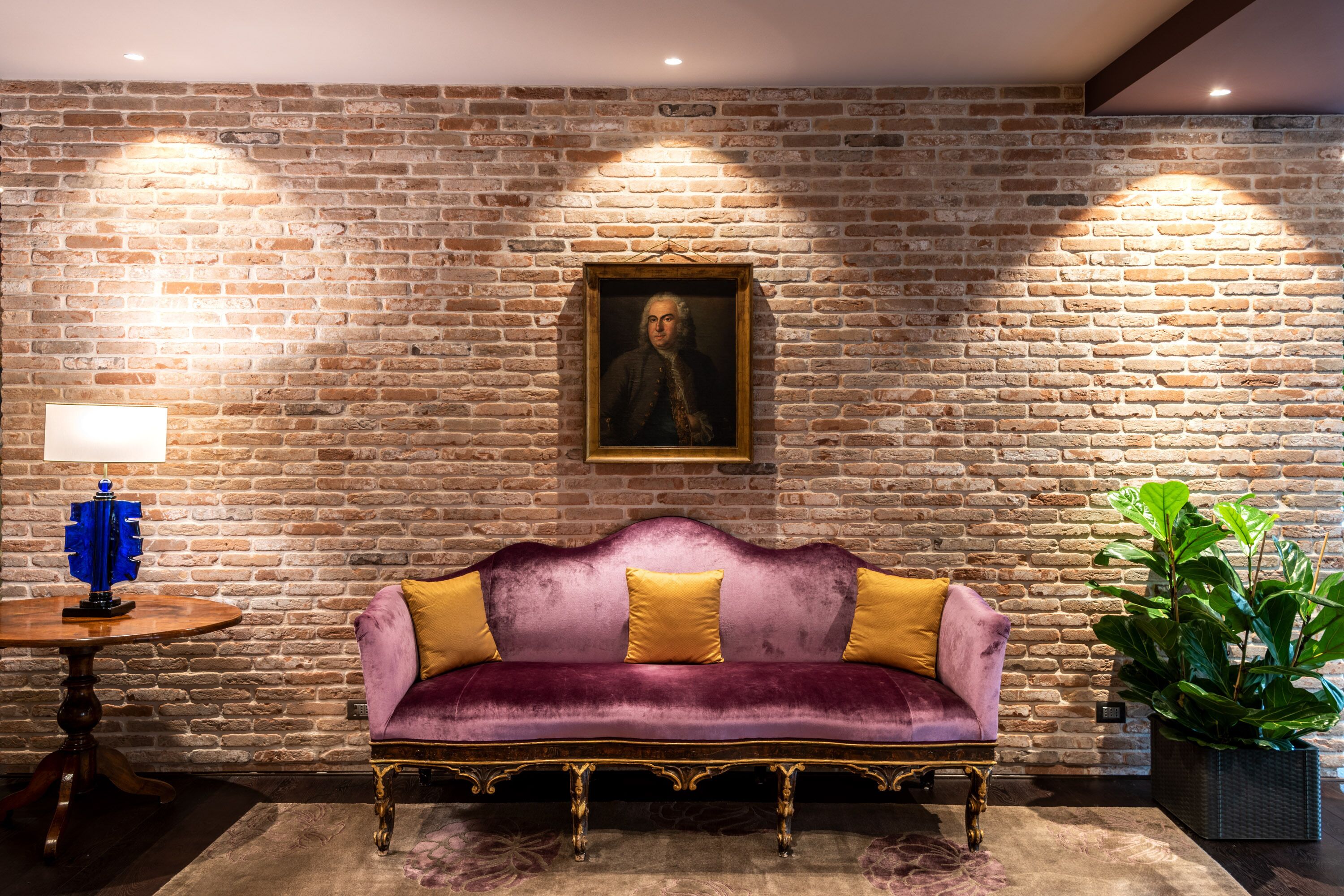 A room with a brick wall, a purple velvet sofa with yellow cushions, a wooden table with a lamp, and a framed portrait above the sofa.