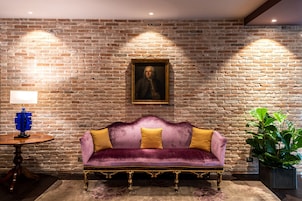 A room with a brick wall, a purple velvet sofa with yellow cushions, a wooden table with a lamp, and a framed portrait above the sofa.