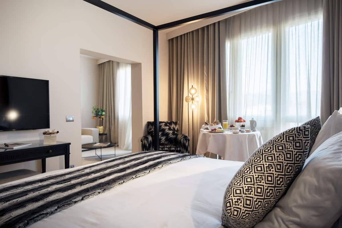 Ein Hotelzimmer mit einem großen Bett, einem Flachbildfernseher, einem kleinen Tisch mit einer Lampe und Blick ins Freie durch das Fenster.