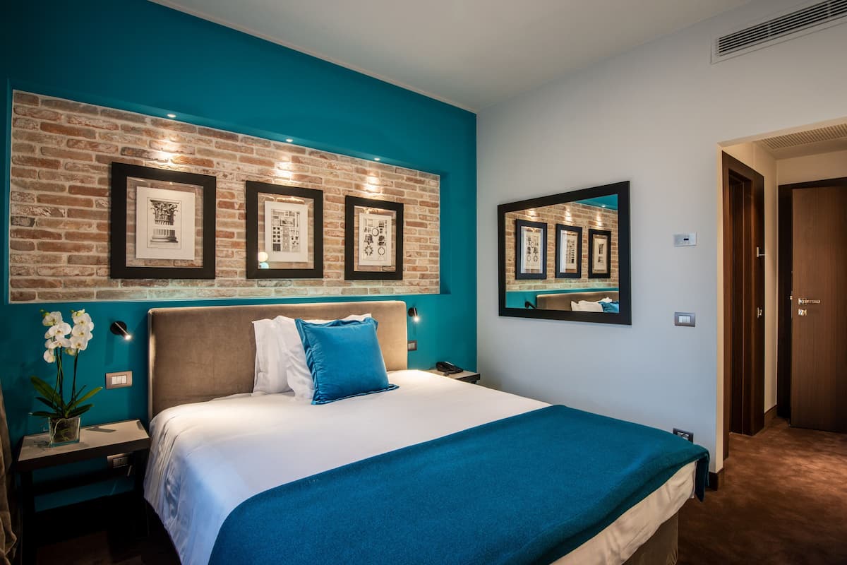 Ein Hotelzimmer mit Backsteinwand, gerahmten Kunstwerken, einem Bett mit blauer Bettwäsche, einem Nachttisch und einer Nachttischlampe sowie einem Spiegel an der Wand.