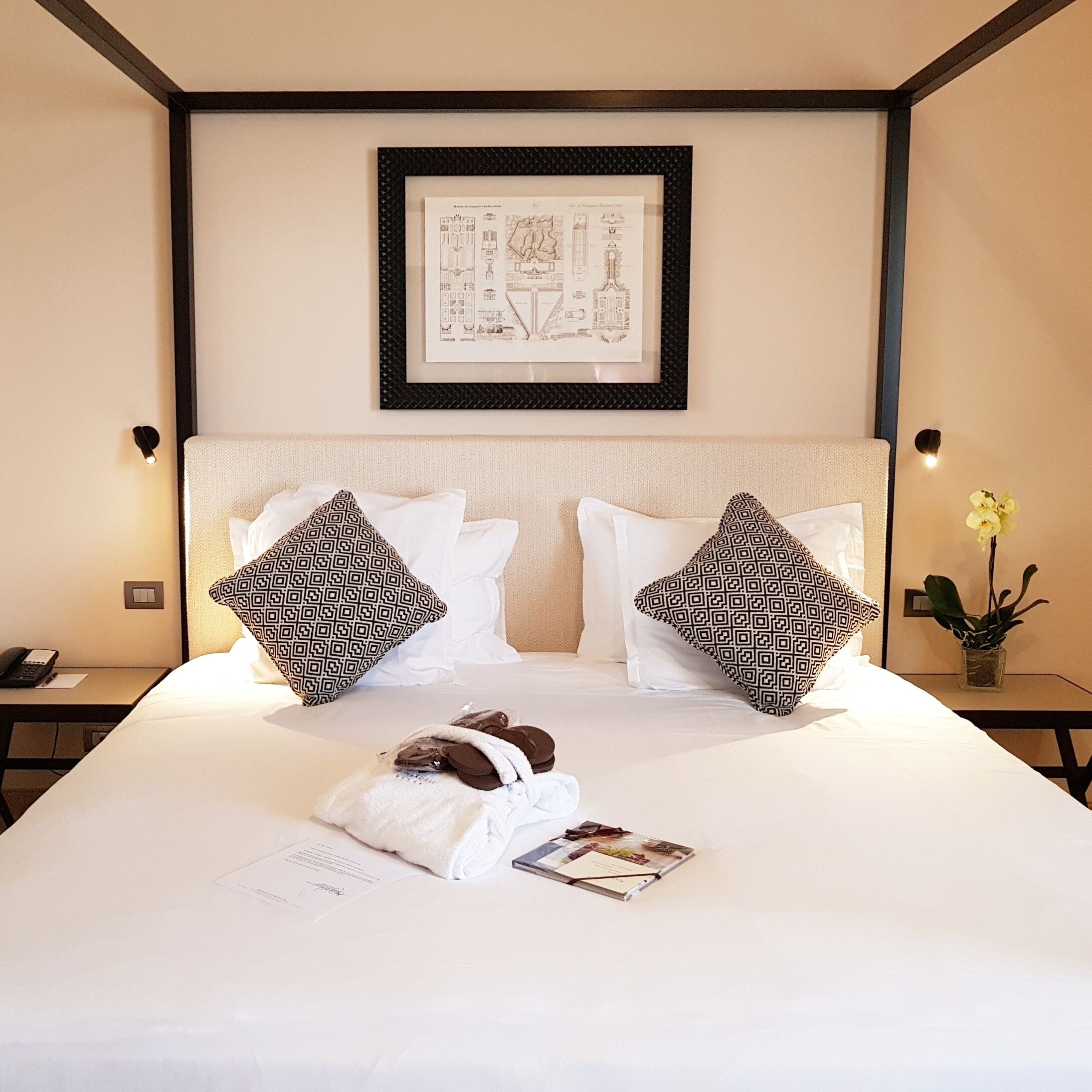 Habitación de hotel con una cama grande, dos almohadas estampadas, un dibujo arquitectónico enmarcado en la pared, una mesita de noche con un teléfono y una planta pequeña.