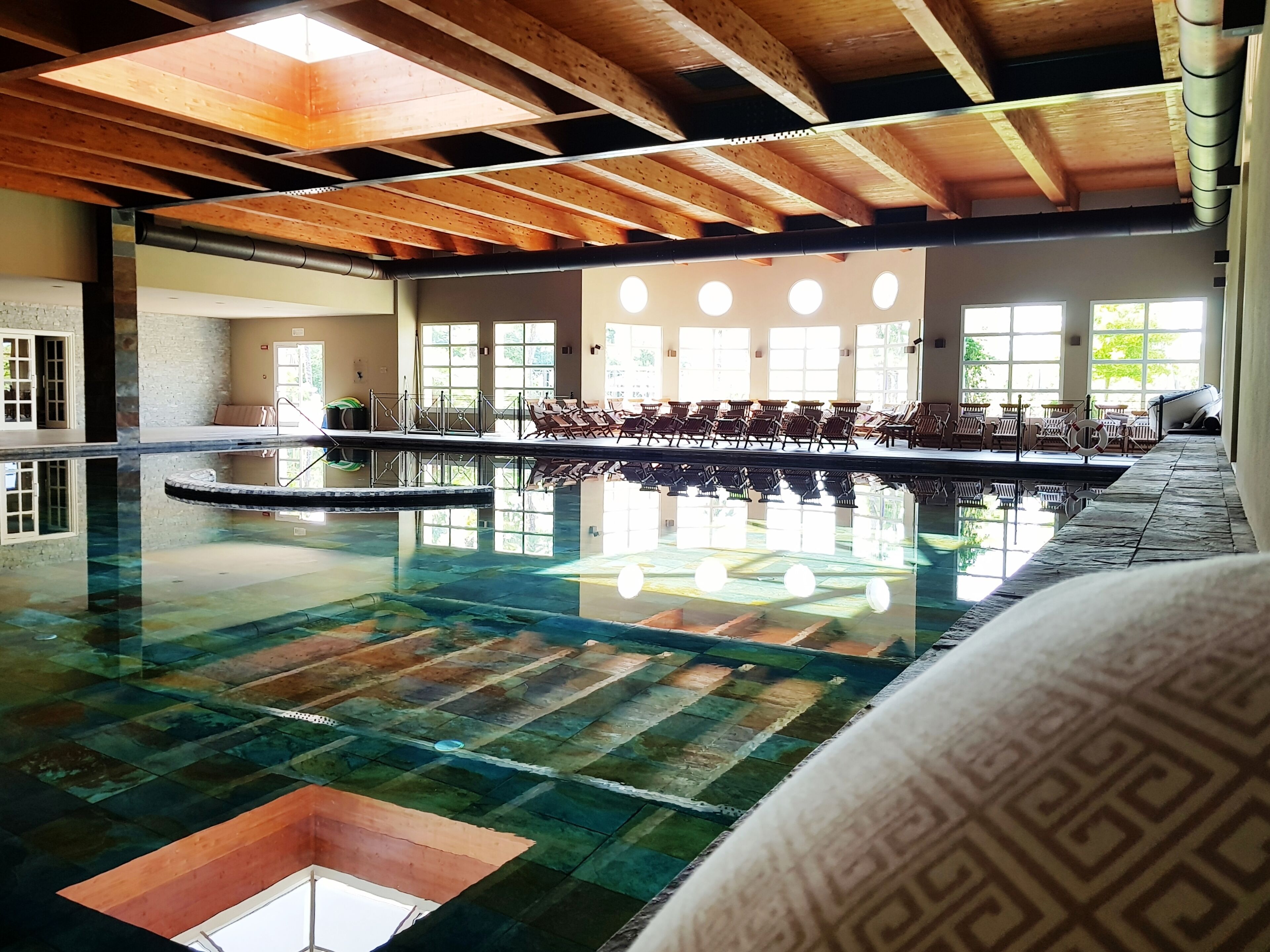 An indoor swimming pool with a wooden ceiling, clear glass walls, and a sunlit area with lounge chairs.