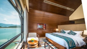 A hotel room with a large window, a bed with white linens, a wooden headboard, a small round table with a bowl of fruit, and a chair. - Ramada by Wyndham Chenzhou Dongjiang Lake (Chenzhou)