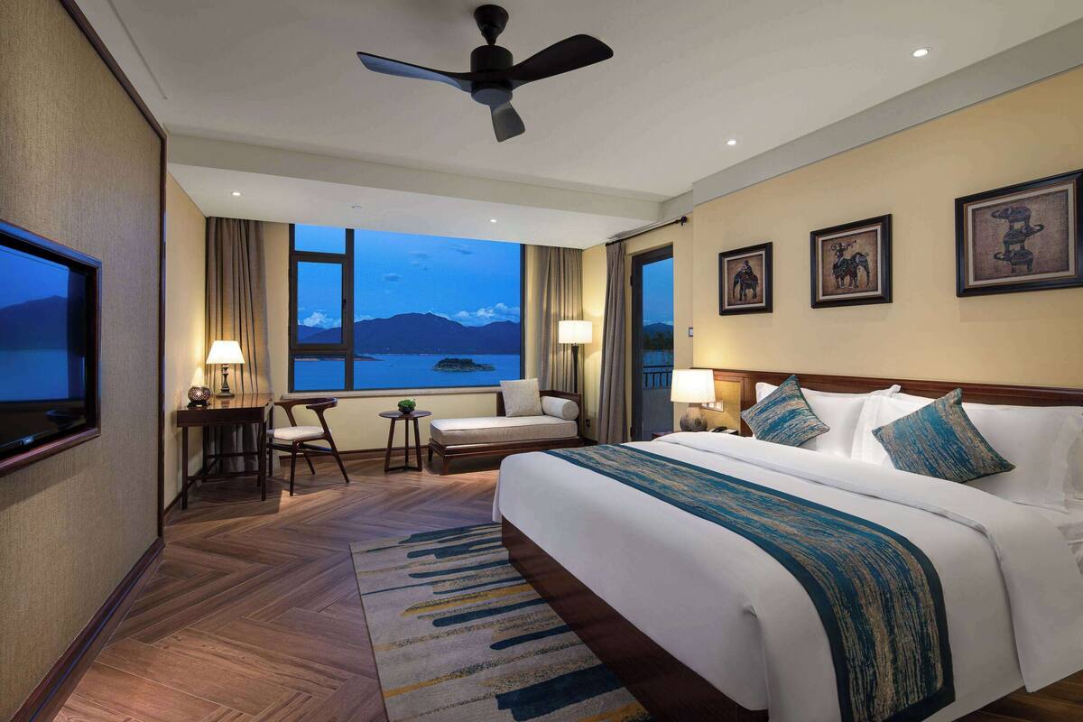Ein Hotelzimmer mit einem großen Bett, einem Schreibtisch, einem Sessel und Blick auf die Berge, sowie einem Deckenventilator.