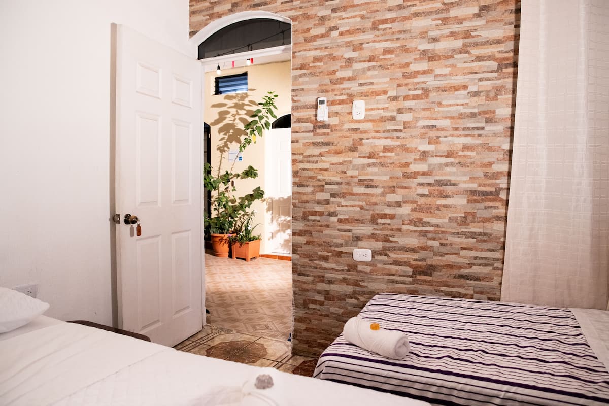 A bedroom with a brick-patterned wall, a bed with a striped blanket, and a white door leading to a hallway with potted plants.