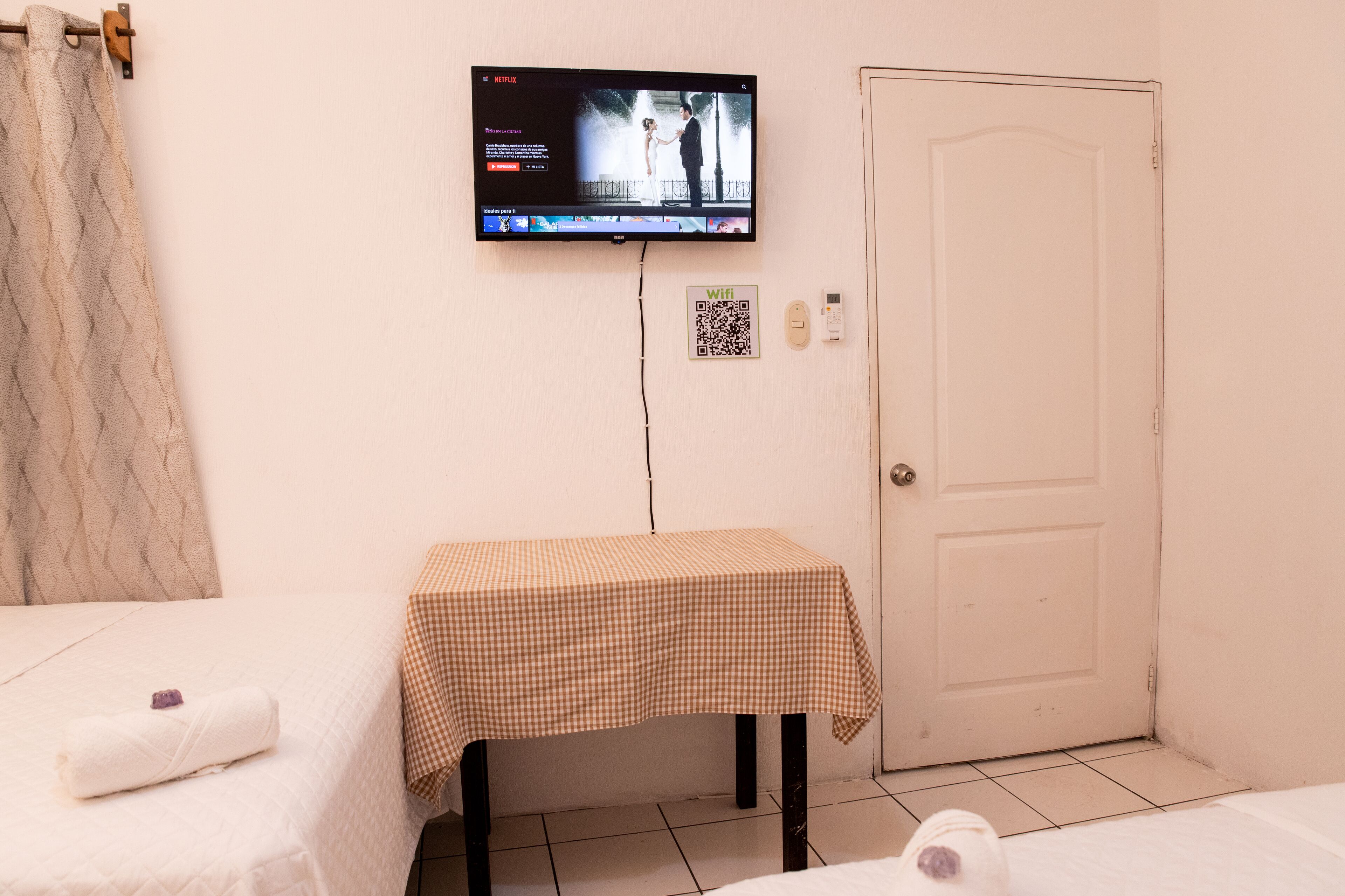 A hotel room with a bed, a small table with a checkered tablecloth, a television mounted on the wall, a door, and a QR code for WiFi.