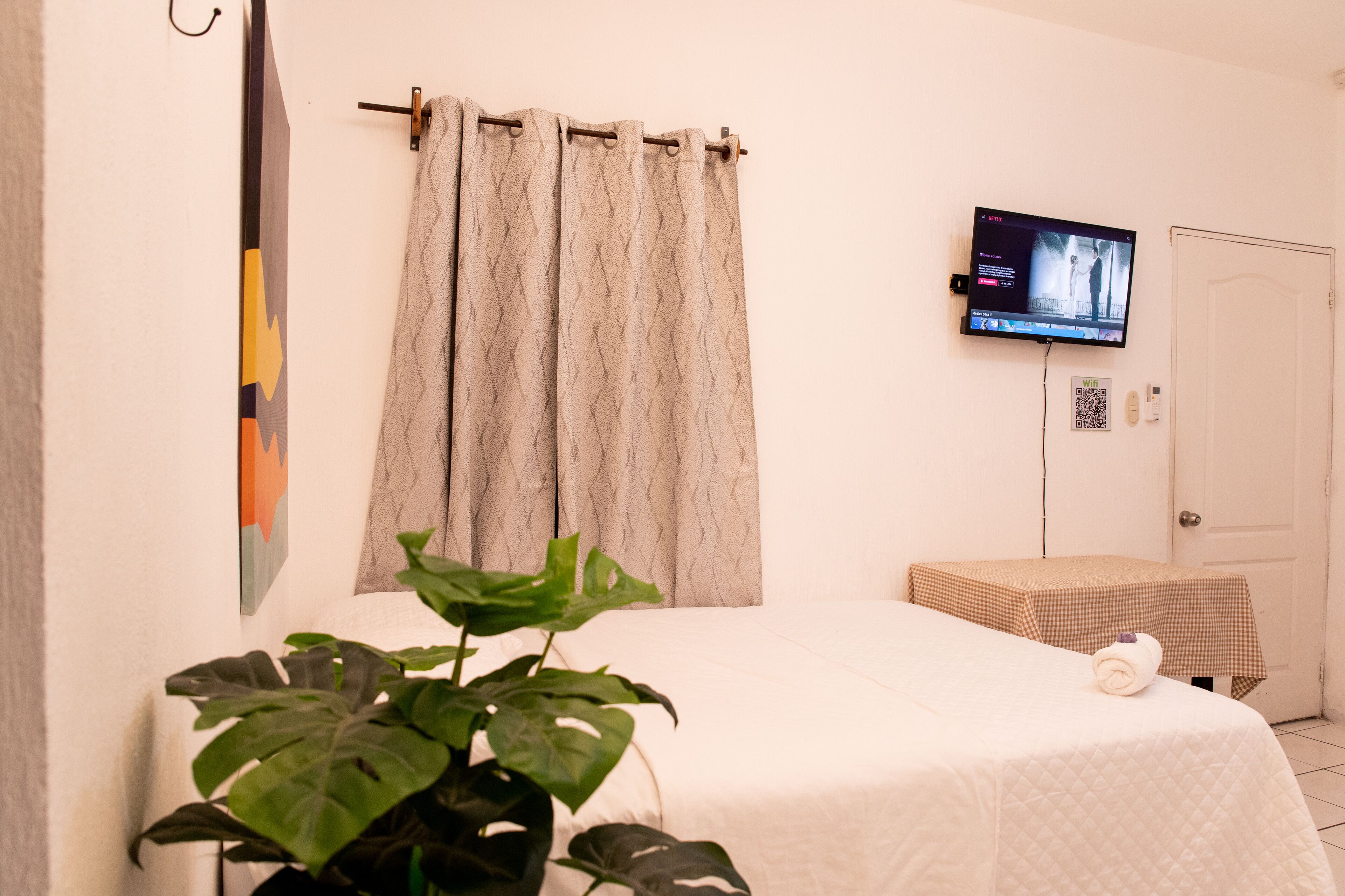 Ein Hotelzimmer mit Bett, Wandfernseher, Vorhang, einem kleinen Tisch und einer Topfpflanze.