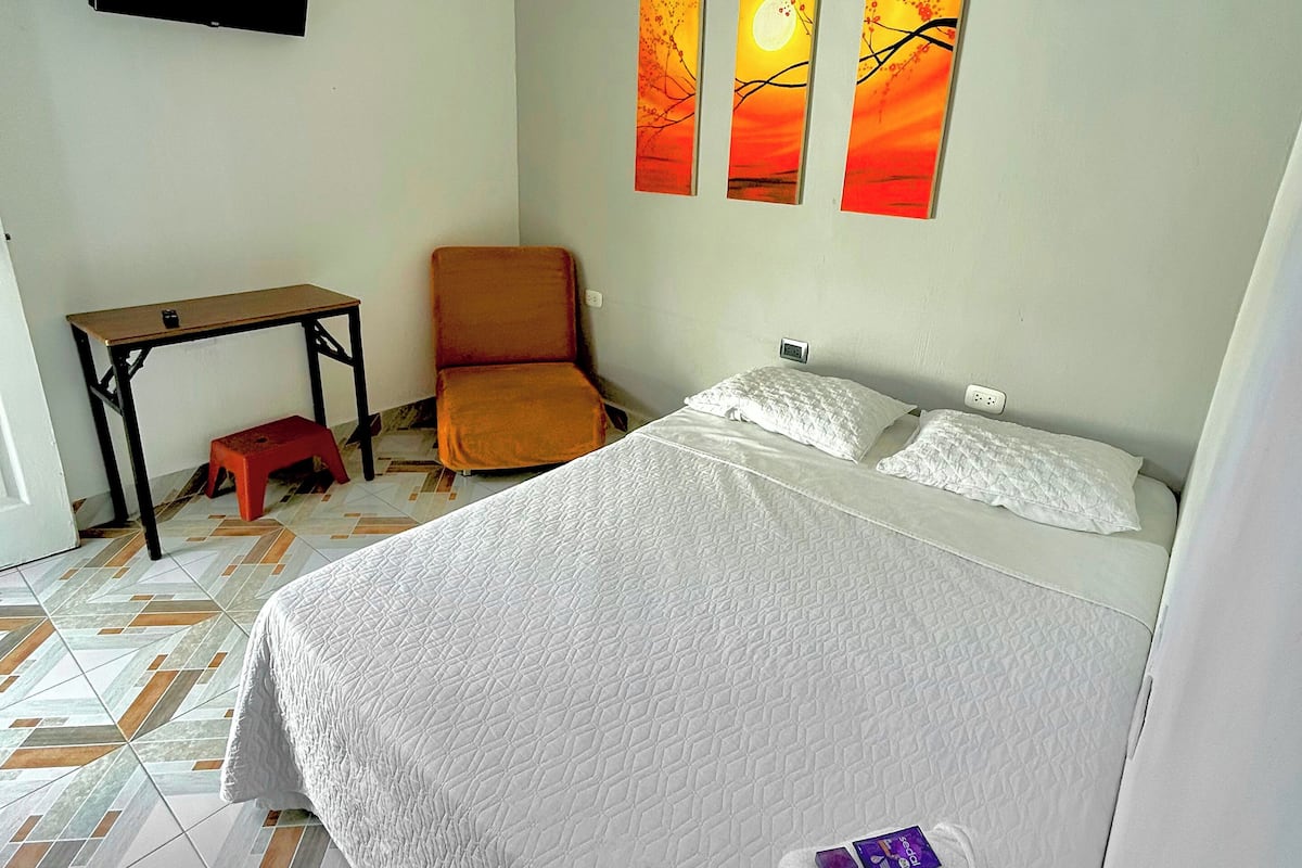 A hotel room with a bed, a chair, a small table, and three paintings on the wall.