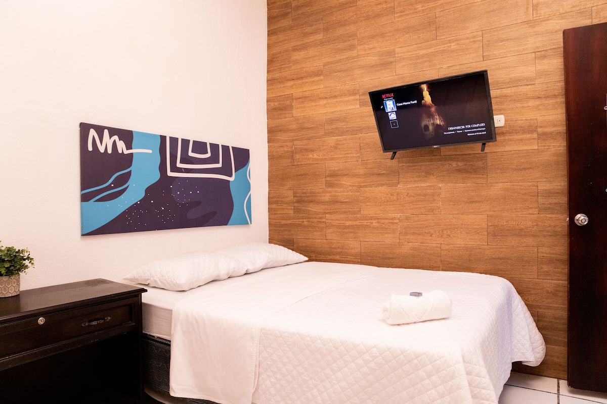 A hotel room with a bed, a wall-mounted TV, a painting, and a wooden floor.