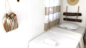Una cama bien hecha con colcha blanca, un bolso a rayas colgado en la pared y un cabecero de madera con un sombrero tejido.