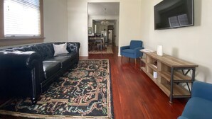 A living room with a black leather sofa, blue armchairs, a wooden coffee table, and a large flat-screen TV mounted on the wall.