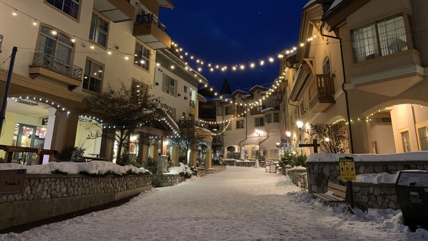 Calle cubierta de nieve con edificios a ambos lados, luces colgantes y un cartel que indica un recorrido por el pueblo.