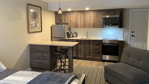 Cocina compacta con armarios de madera, refrigerador, microondas y estufa. Hay un taburete de bar y un sofá gris en la sala.