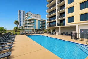Ein Swimmingpool mit Liegestühlen, ein mehrstöckiges Gebäude mit Balkonen und Palmen.