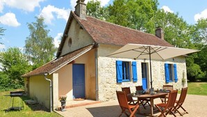 A rustic stone house with a tiled roof, a covered patio with wooden furniture, and a barbecue grill.
