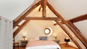 A bedroom with a sloped ceiling, wooden beams, a round skylight, a bed with pillows, two bedside tables with lamps, and a chair.