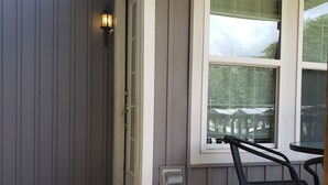 Un portico riparato con pavimento in legno, una sedia in metallo e una finestra che si affaccia su una casa e alberi.