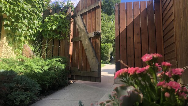 A wooden gate leading to a driveway, surrounded by greenery and a clear sky.