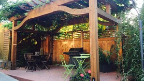 A wooden pergola with a grill and chairs, surrounded by plants and a wooden fence.