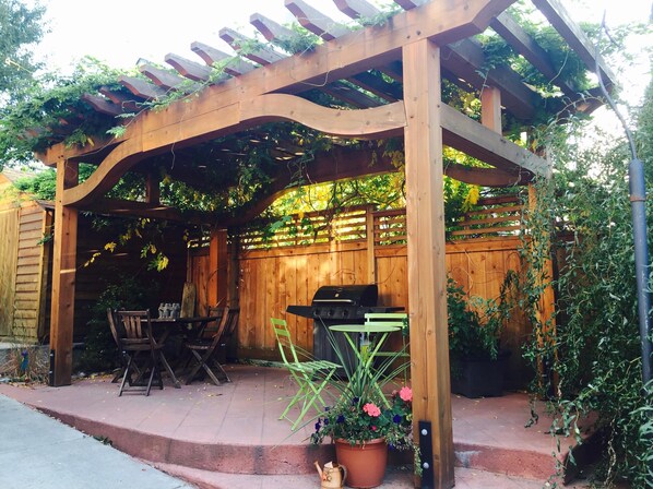 Una pérgola de madera con una parrilla y sillas, rodeada de plantas y un cerco de madera.