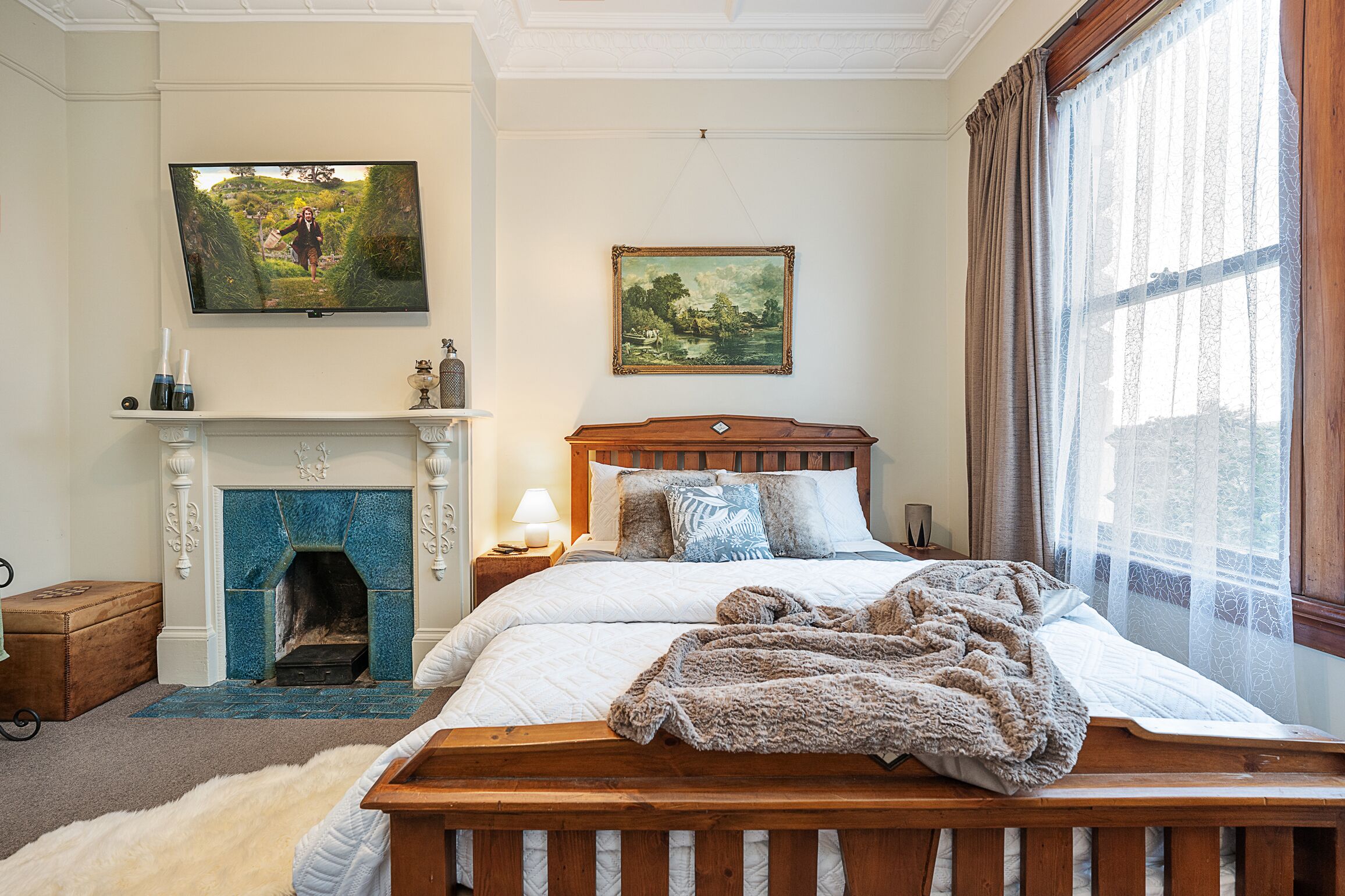 A bedroom with a fireplace, a bed with a blanket, a TV, and a painting on the wall.