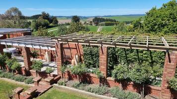 Uma pérgola de tijolos com uma mesa e cadeiras por baixo, rodeada por um jardim bem cuidado com várias plantas e gramado.