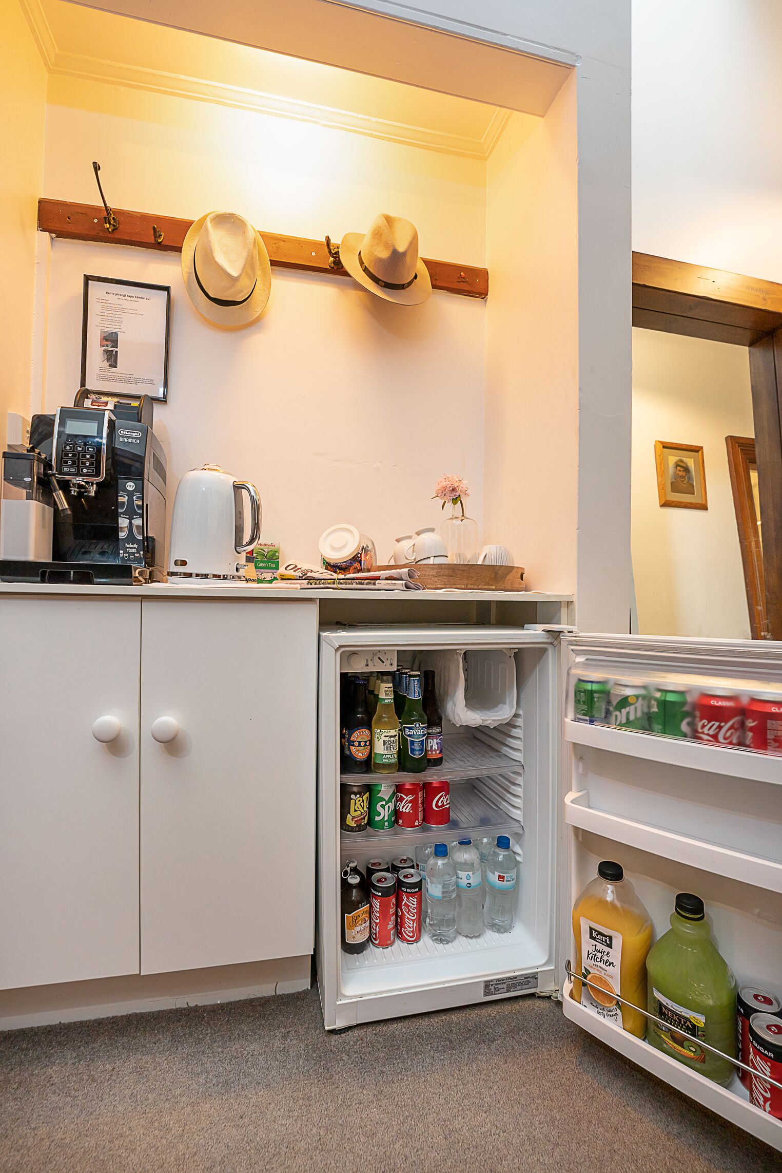 Una cocina con un refrigerador lleno de bebidas, una cafetera y sombreros en la pared.