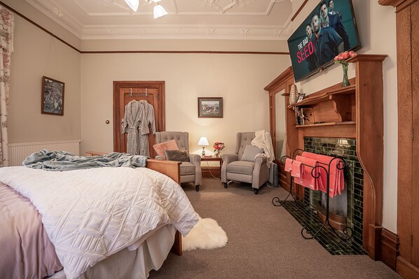 Un dormitorio acogedor con una cama grande, dos sillones, una mesita de noche con una lámpara y una chimenea con un perchero rojo.