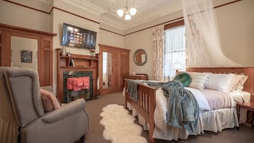 Un dormitorio con cama, sillón, chimenea, televisor y un amplio ventanal con cortinas.