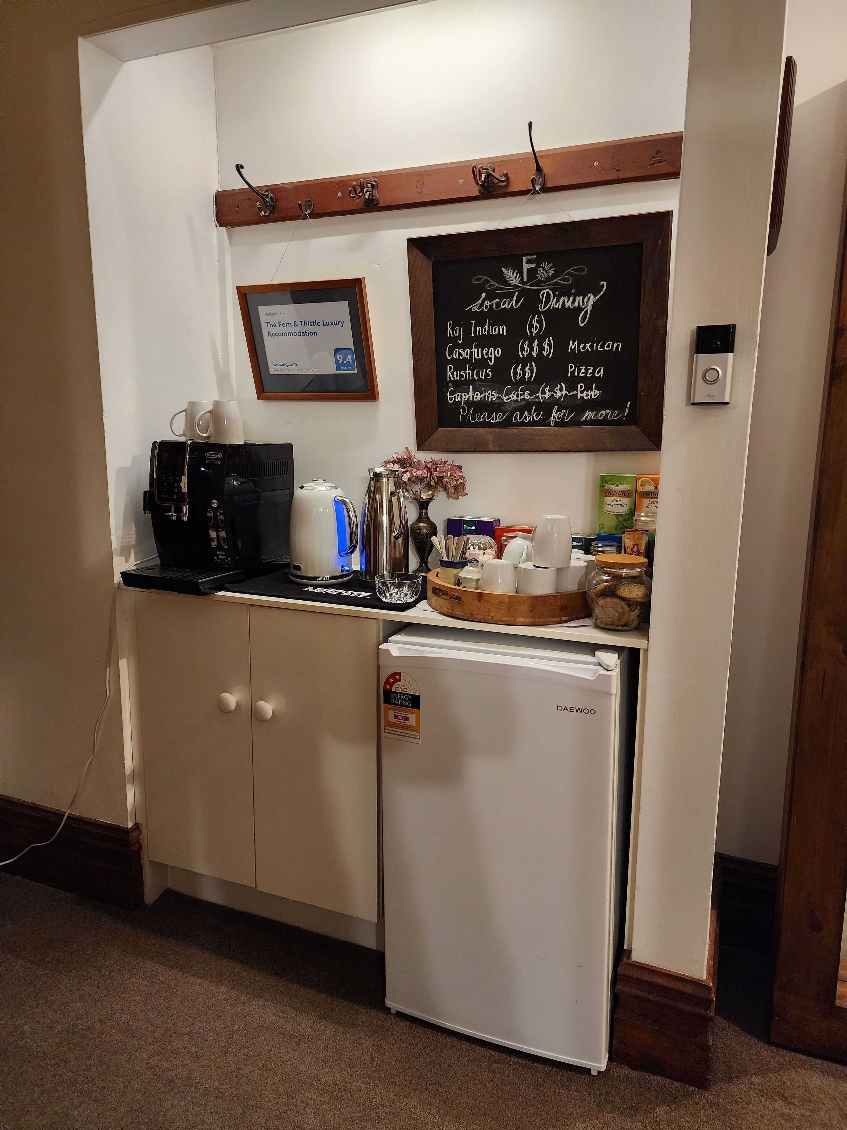 Une cuisine compacte équipée d’une machine à café, d’un réfrigérateur et d’un tableau noir affichant une liste des restaurants locaux.