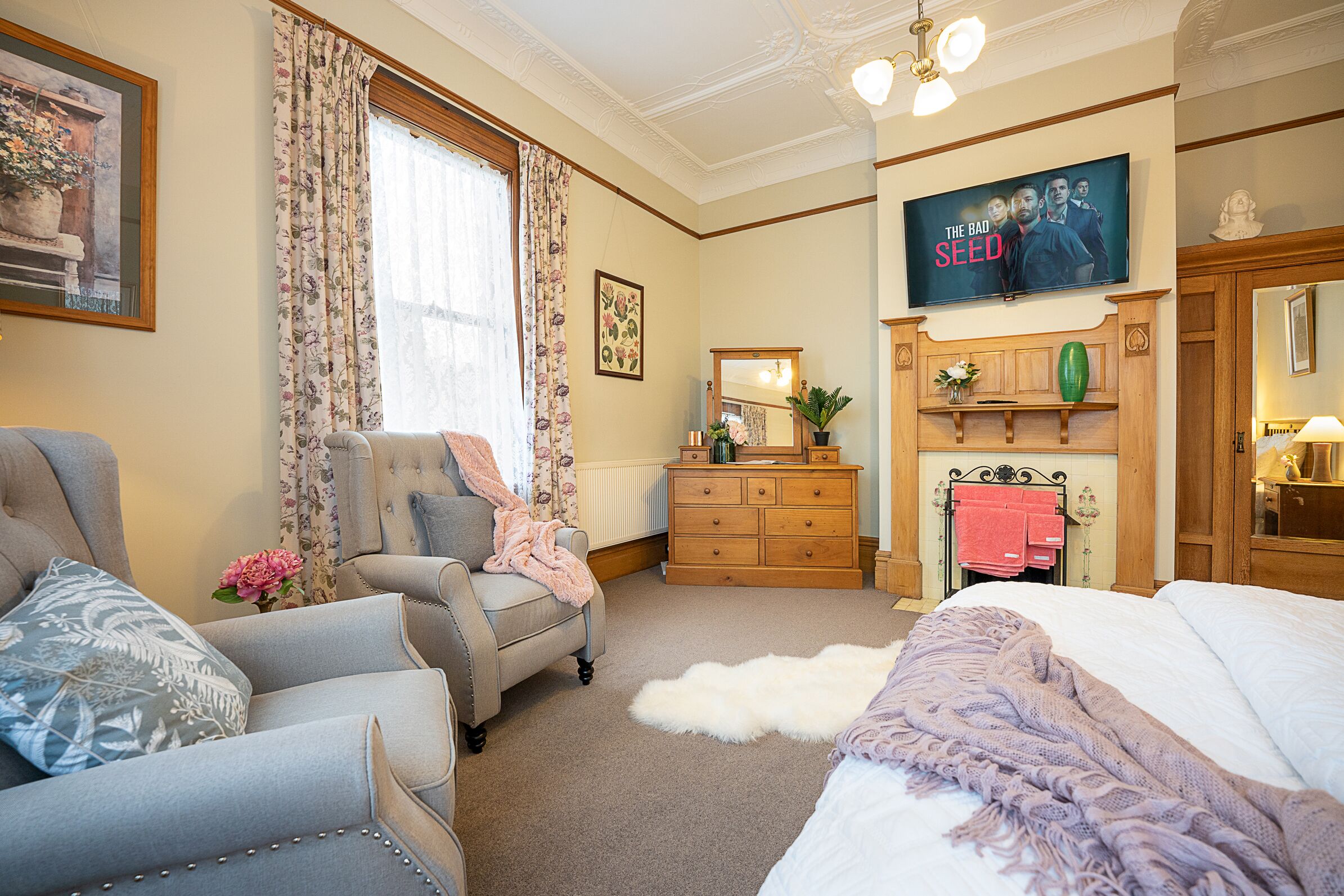 A well-furnished bedroom with a bed, two armchairs, a dresser, and a television displaying 'The Bad Seed'.