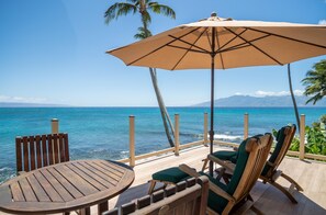 A wooden deck with a table and chairs, a large umbrella, and a view of the ocean and mountains. - Oceanfront, hot tub, pool, perfect location! (Lahaina)
