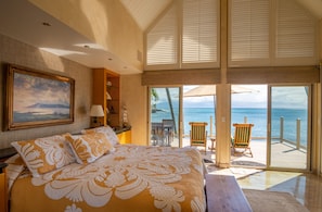 A bedroom with a large bed, a painting, a nightstand, and a view of the ocean through a sliding door. - Oceanfront, hot tub, pool, perfect location! (Lahaina)