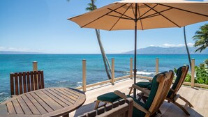 A wooden deck with a table and chairs, an umbrella, and a view of the ocean and mountains. - 3br/3.5ba Oceanfront home with pool and hot tub! (Lahaina)