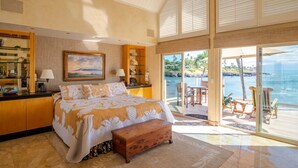 A bedroom with a large bed, a bench, a desk with a lamp, a painting, and a view of the ocean through a sliding door. - 3br/3.5ba Oceanfront home with pool and hot tub! (Lahaina)