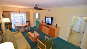 A hotel room with a living area, a dining table with chairs, a television, a ceiling fan, and a bathroom visible through an open door.