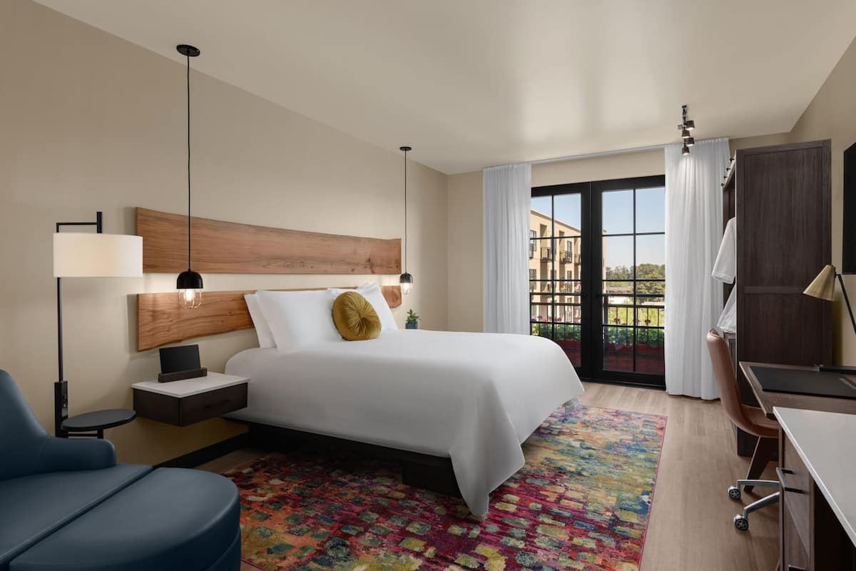 A modern hotel room with a large bed, a desk, a chair, a desk lamp, a floor lamp, and a view of the outside.