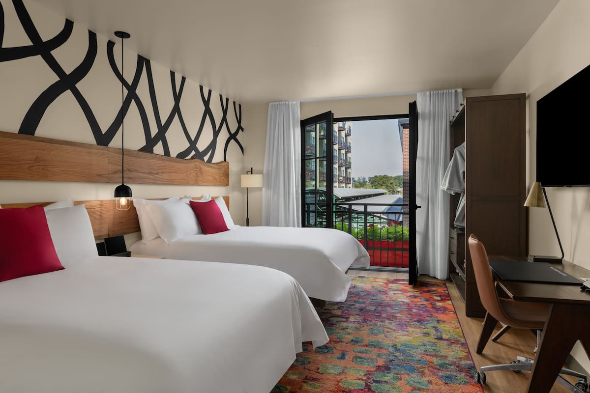 A hotel room with two beds, a desk, a chair, a colorful carpet, and a view of the city.