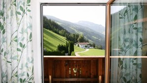 Una habitación con piso de madera, vista a un paisaje verde con colinas y una casa, además de un banco de madera.