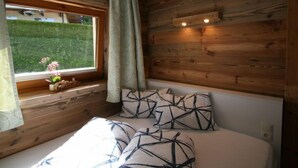 Un dormitorio con paredes de madera, un ventanal con vistas a la vegetación y una cama con almohadones estampados.