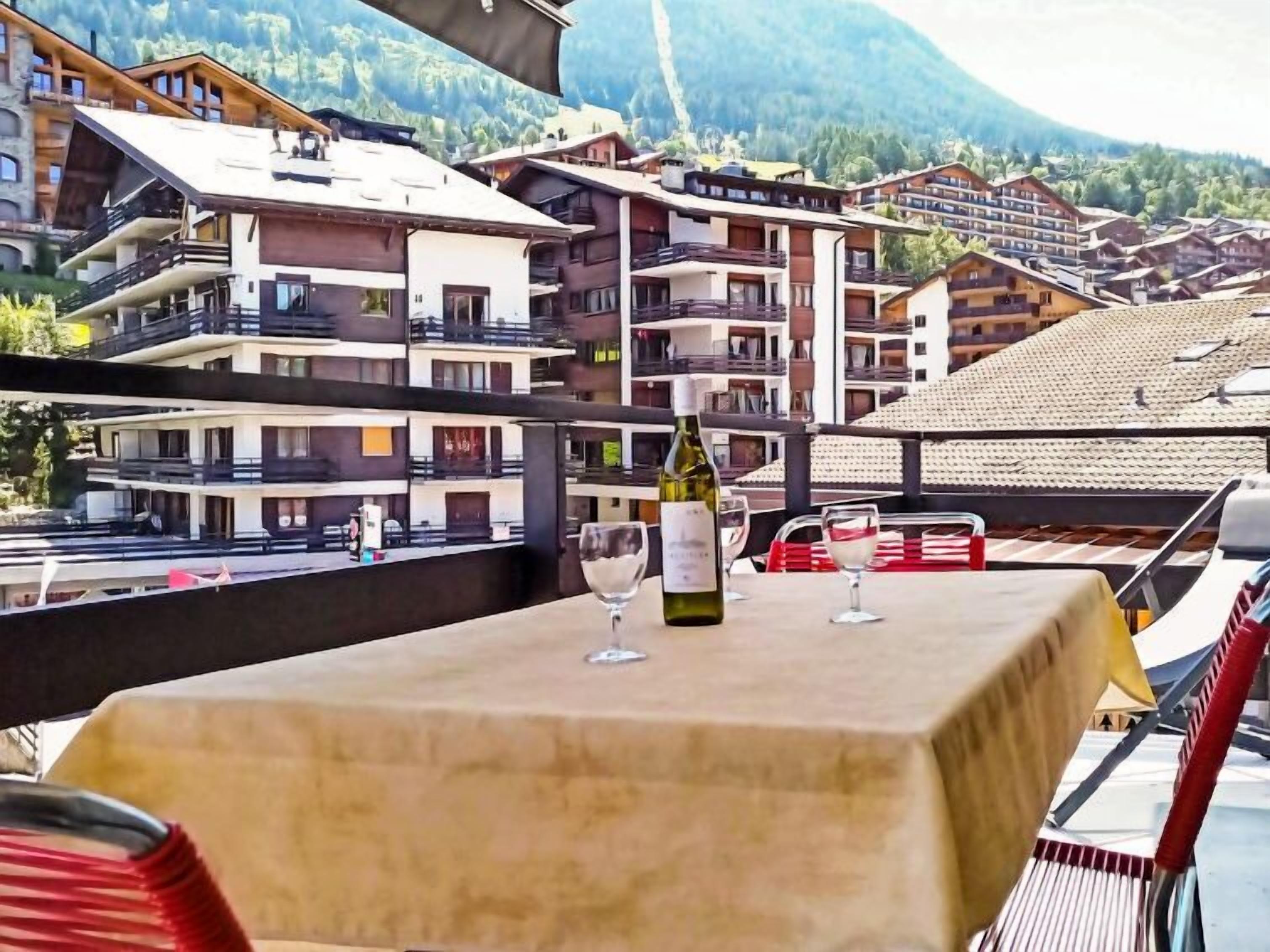 Uma mesa com uma garrafa de vinho e duas taças, com vista para uma aldeia montanhosa com edifícios tradicionais.