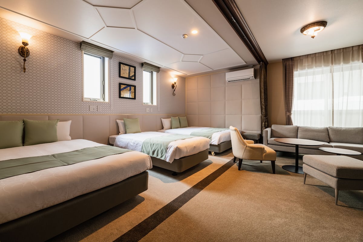 A hotel room with three beds, a sofa, a small table, and a chair. There are wall-mounted lights and a window with curtains.