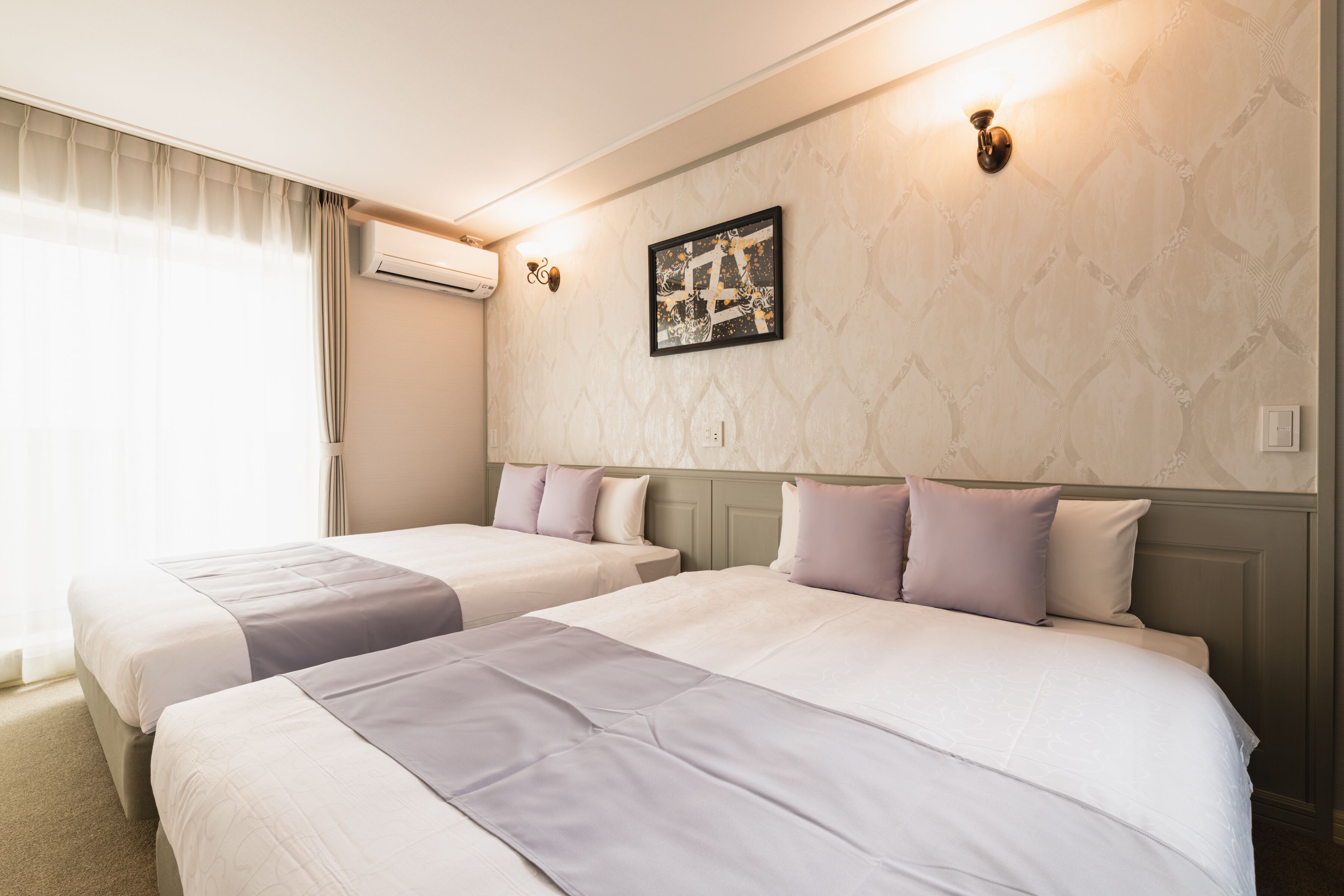 Habitación de hotel con dos camas, un ventanal con cortinas, un equipo de aire acondicionado y una lámpara fijada a la pared.