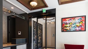 A reception area with a check-in counter, a glass door entrance, a red chair, and a colorful abstract painting on the wall. - Grand Base Hakata-Ⅰ (Fukuoka)