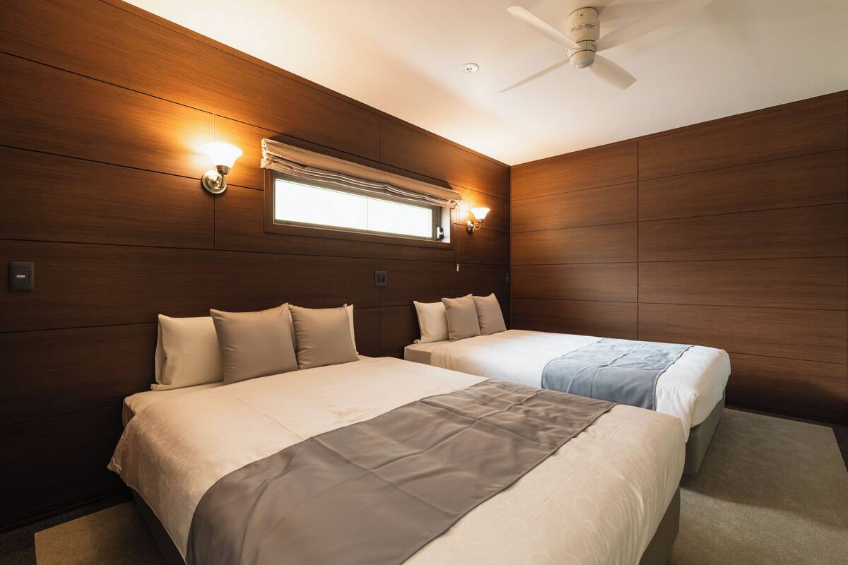 A hotel room with two beds, wooden panel walls, a ceiling fan, and a window with blinds.