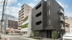 Un edificio moderno con fachada oscura, varios balcones y vista a la calle con coches y vegetación.