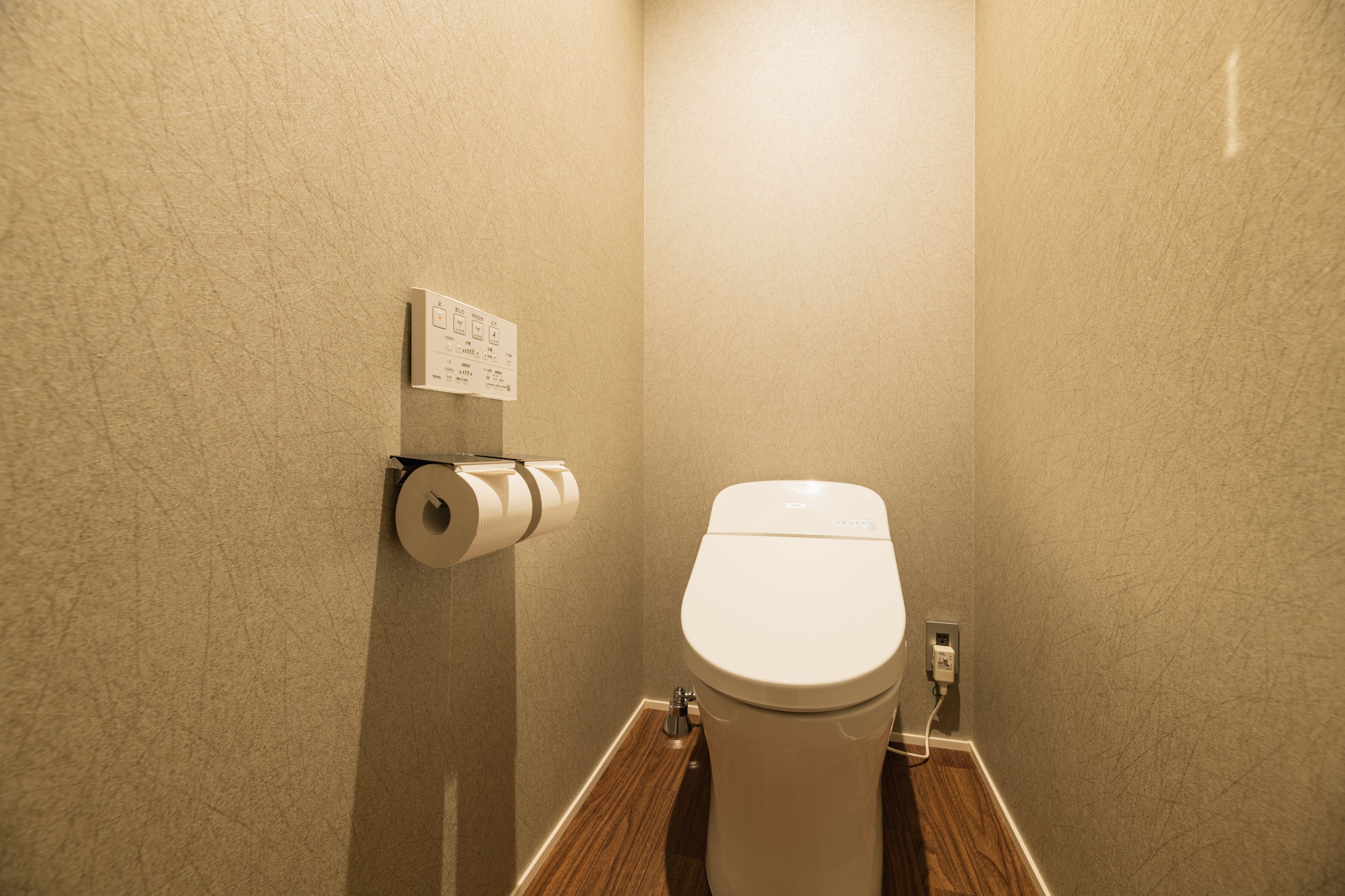 A small bathroom with a toilet, a paper towel dispenser, and a wall-mounted electrical outlet.