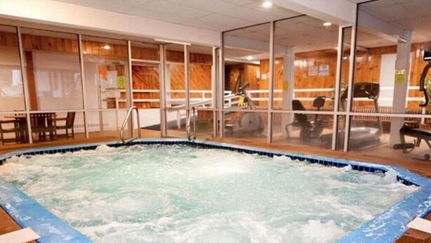Ein Whirlpool mit umlaufender Holzterrasse, ein Indoor-Sitzbereich mit Tischen und Stühlen sowie Trainingsgeräte im Hintergrund.