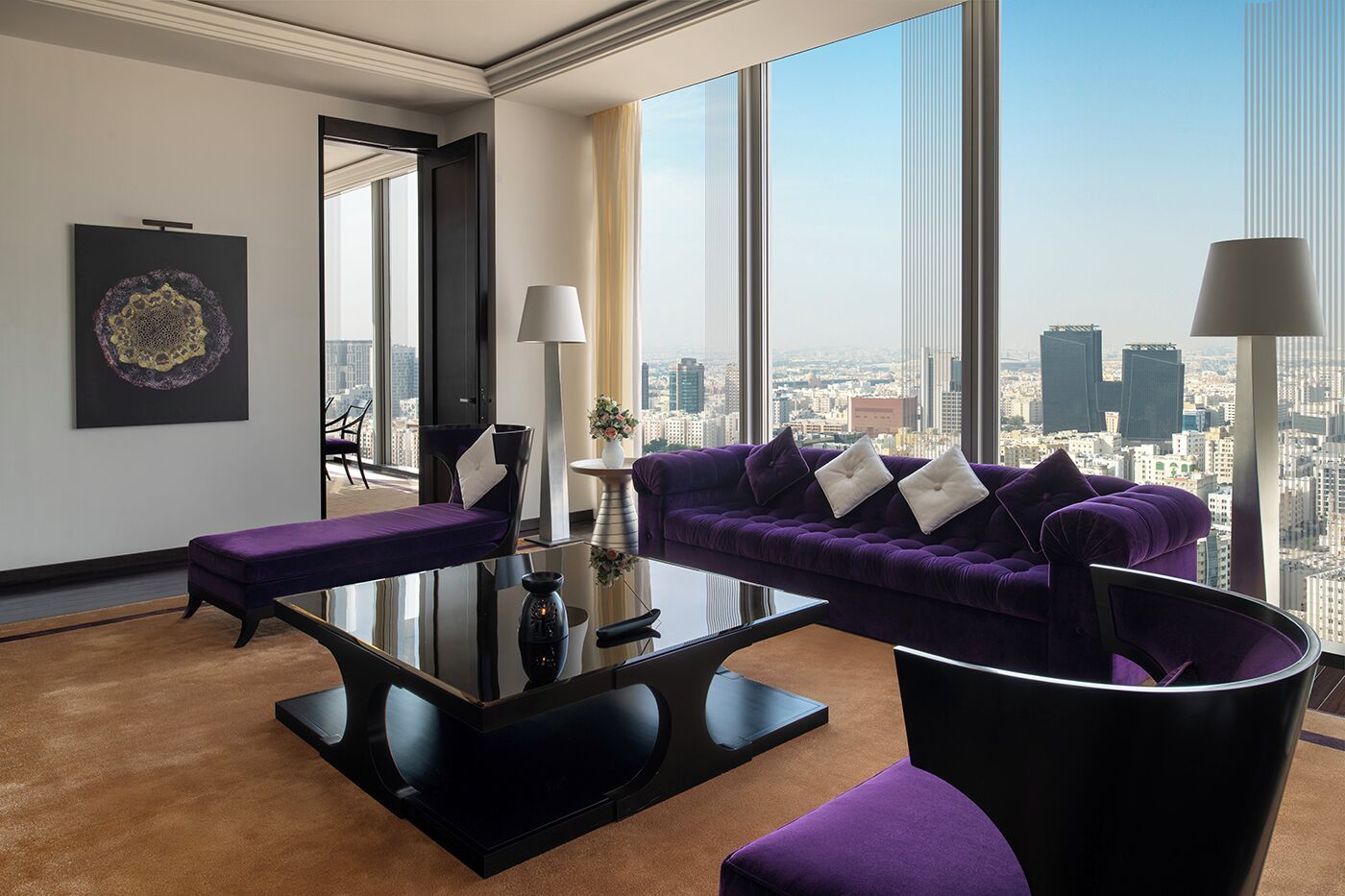 A modern hotel room with a large city view, a purple sofa, a glass coffee table, and a black armchair.