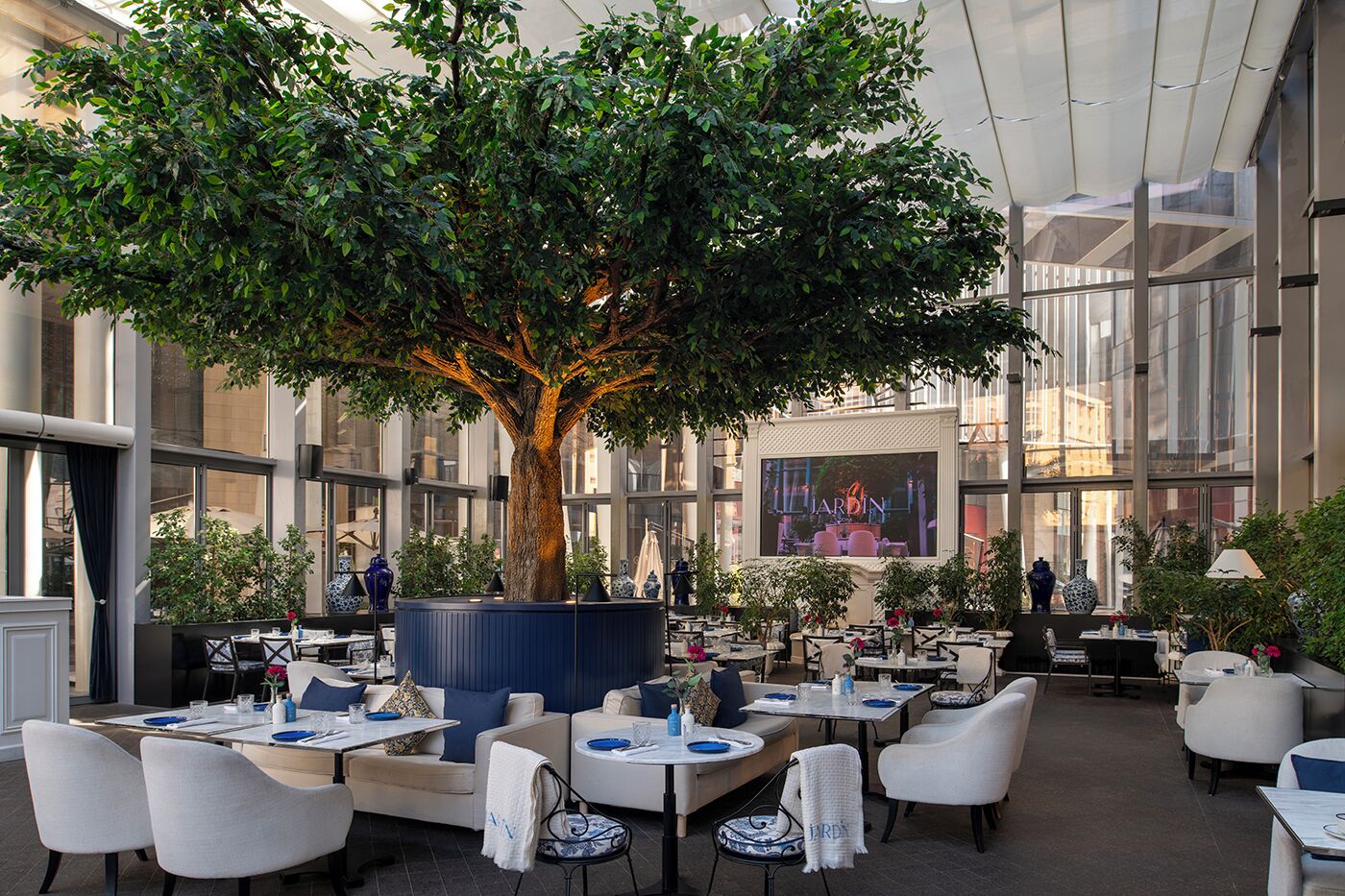 An indoor dining area with tables, chairs, and a large tree in the center.